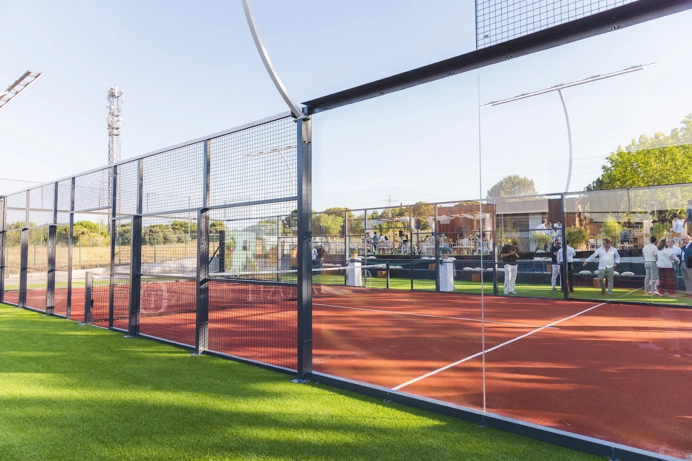 Wide photograph of Tizona’s outdoor red padel court with its courtside social area, trees and wood-finished buildings